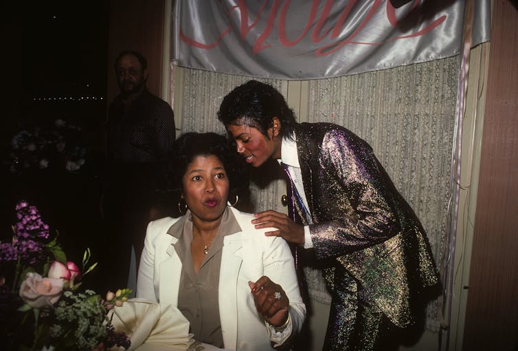 Katherine Jackson, dressed in a white jacket over an olive green shirt, sits at a table covered in flowers; her son Michael Jackson stands behind her, wearing a shiny suit.